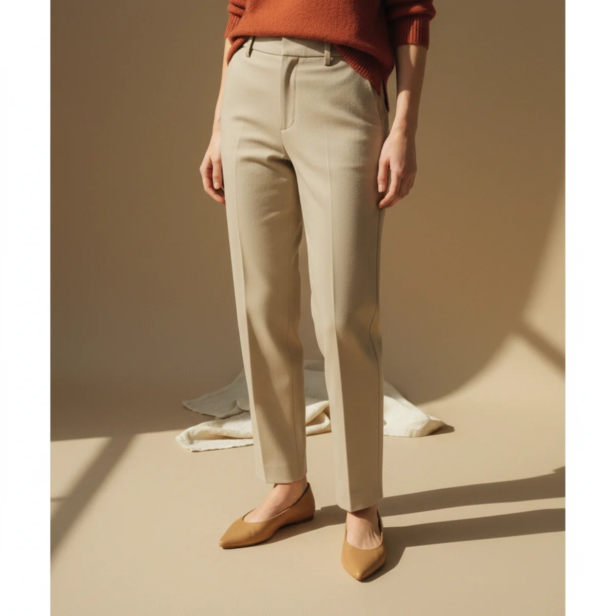 Faceless waist-down shot of beige straight-leg trousers with tan pointed-toe flat shoes and the hem of a rust-orange knit top