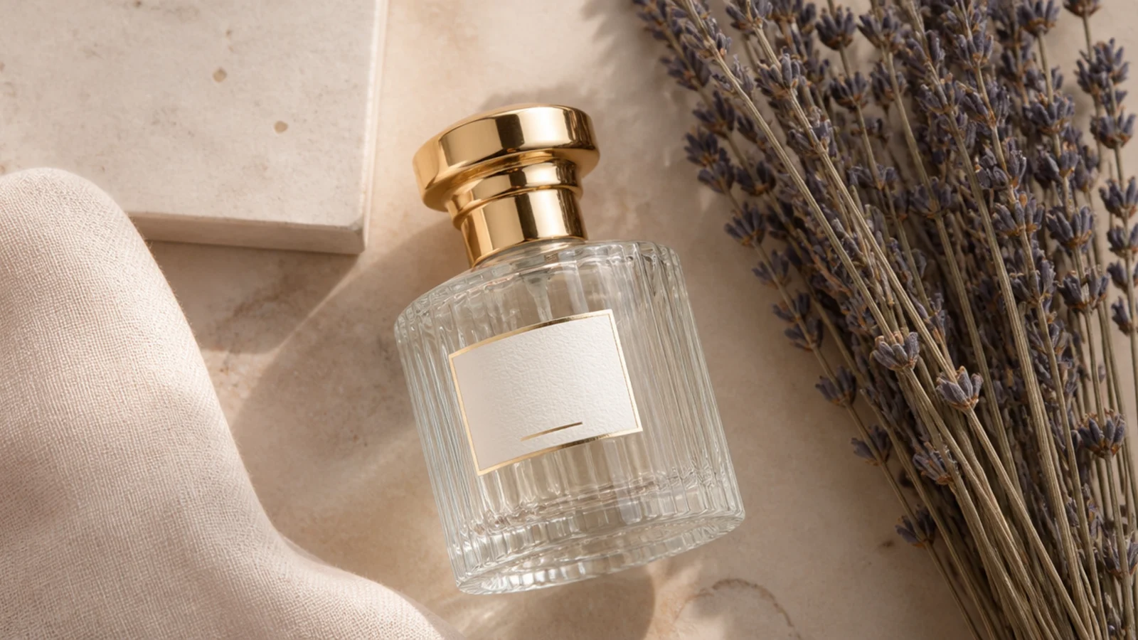 Flat-lay of an elegant clear glass perfume bottle with a gold cap on a warm neutral surface beside dried lavender