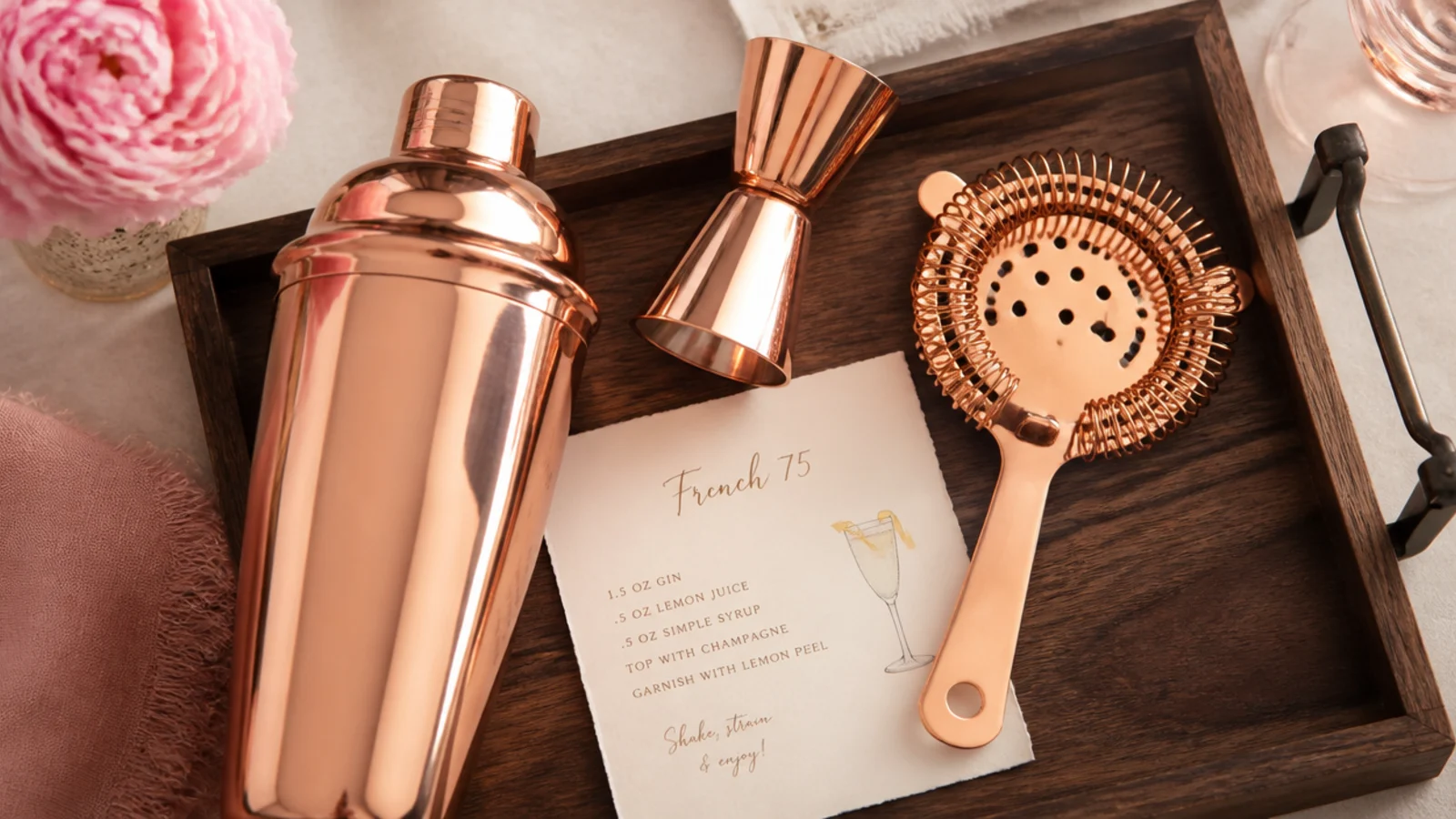 Copper cocktail shaker, jigger and strainer arranged on a dark wood tray with a recipe card