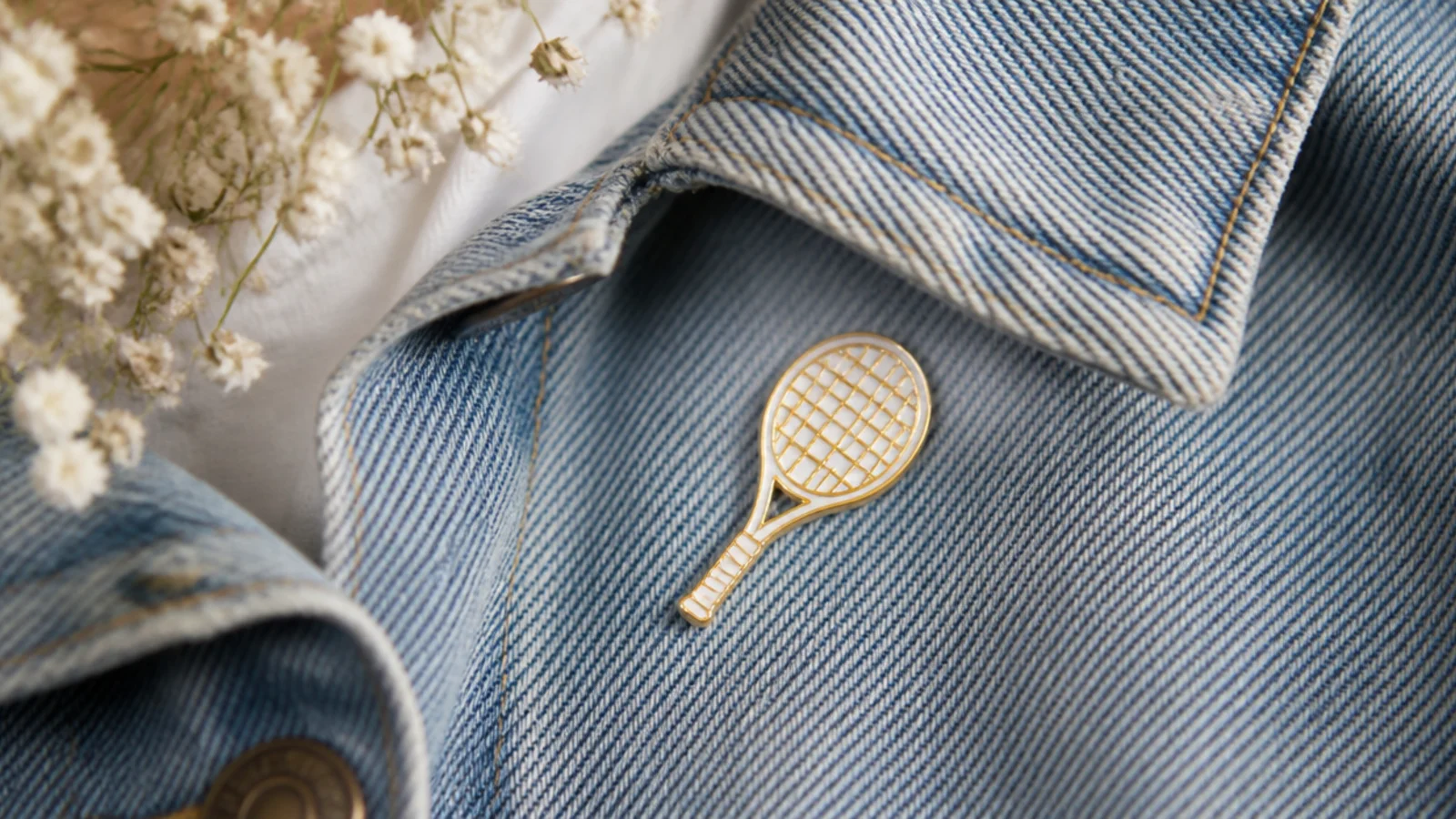 Faceless close-up of a small gold enamel pin shaped like a tennis racket on a denim jacket