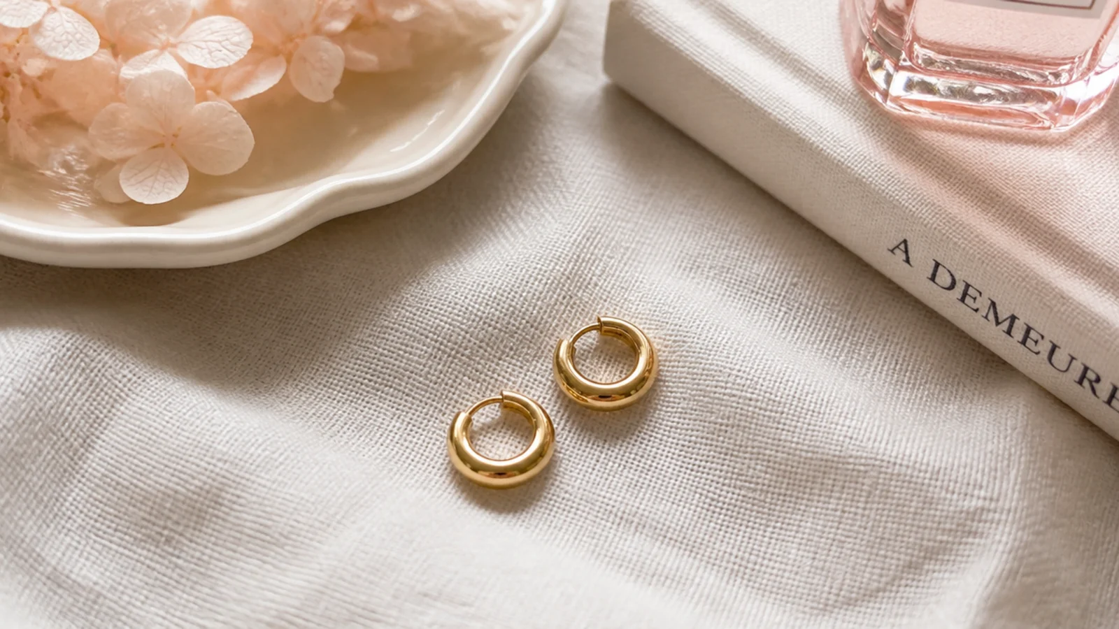 Faceless close-up of small gold-plated huggie hoop earrings on a linen surface