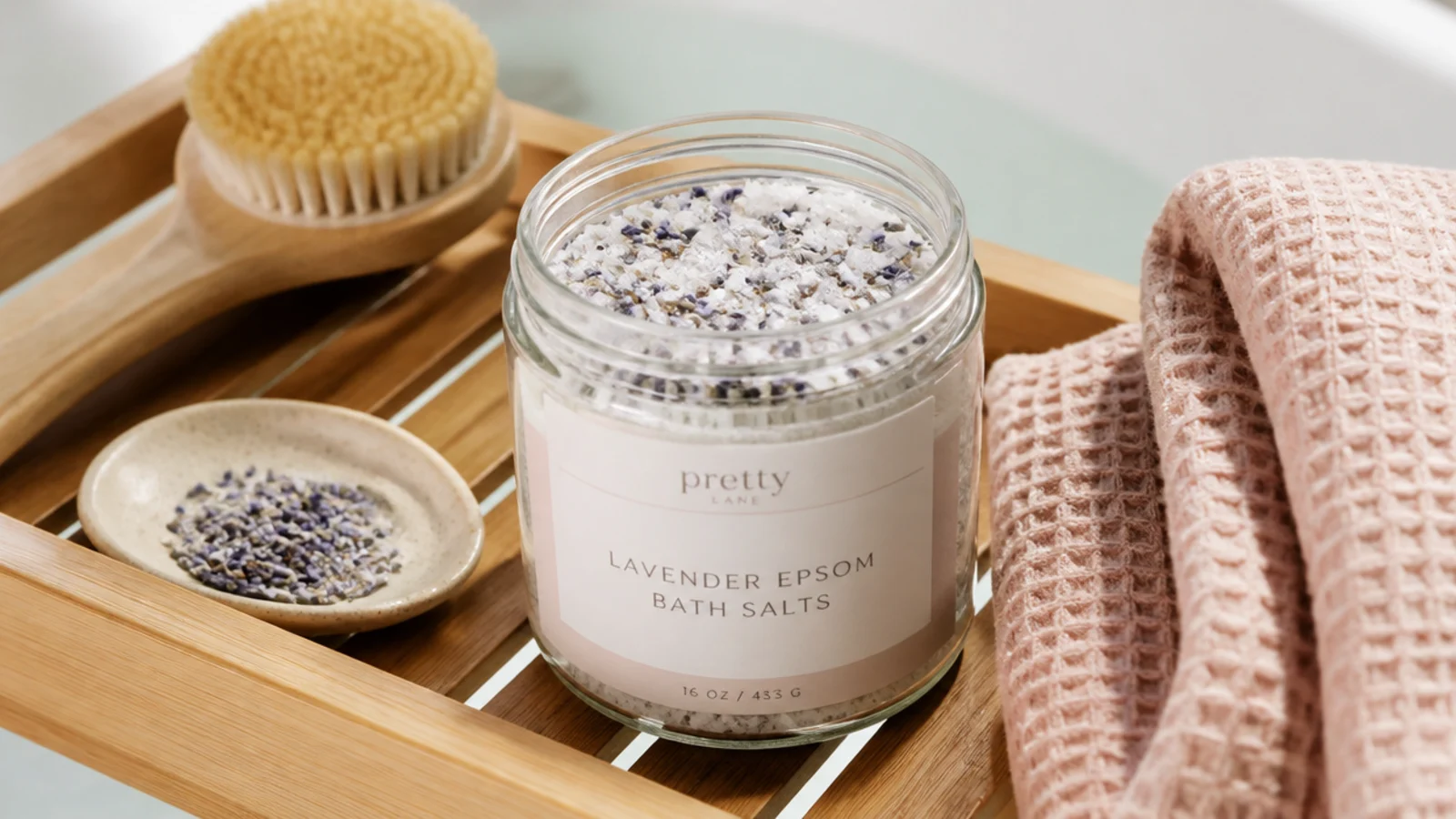 Faceless flat-lay of a glass jar of lavender Epsom bath salts with dried buds on a bath tray