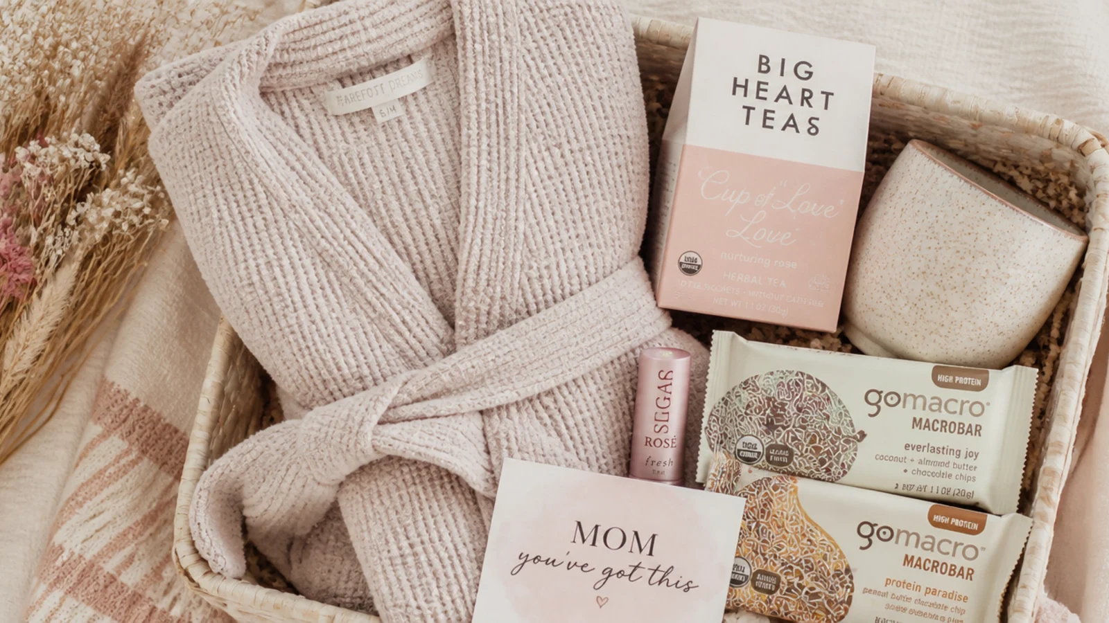 Faceless overhead shot of a new mom basket with a soft robe, lip balm, snack bars and tea