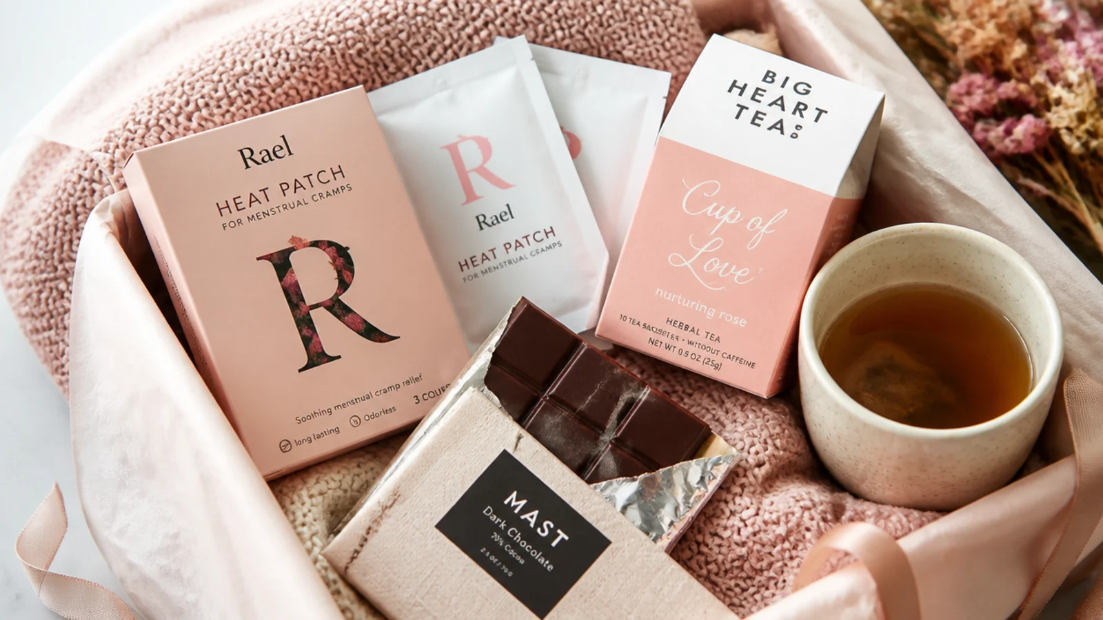 Faceless flat lay of a period-friendly recovery basket with heat patches, dark chocolate and ginger tea