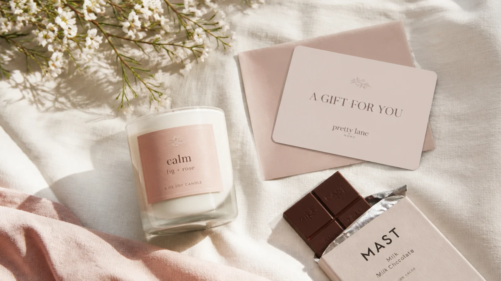 Faceless flat-lay of neutral safe gifts - a candle, chocolate and a gift card - on cream linen