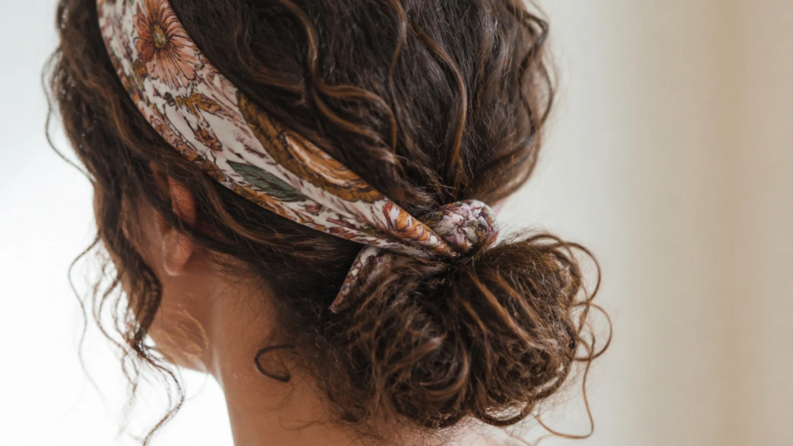 Low curly bun at the nape with a printed silk scarf tied as a headband over frizzy curls
