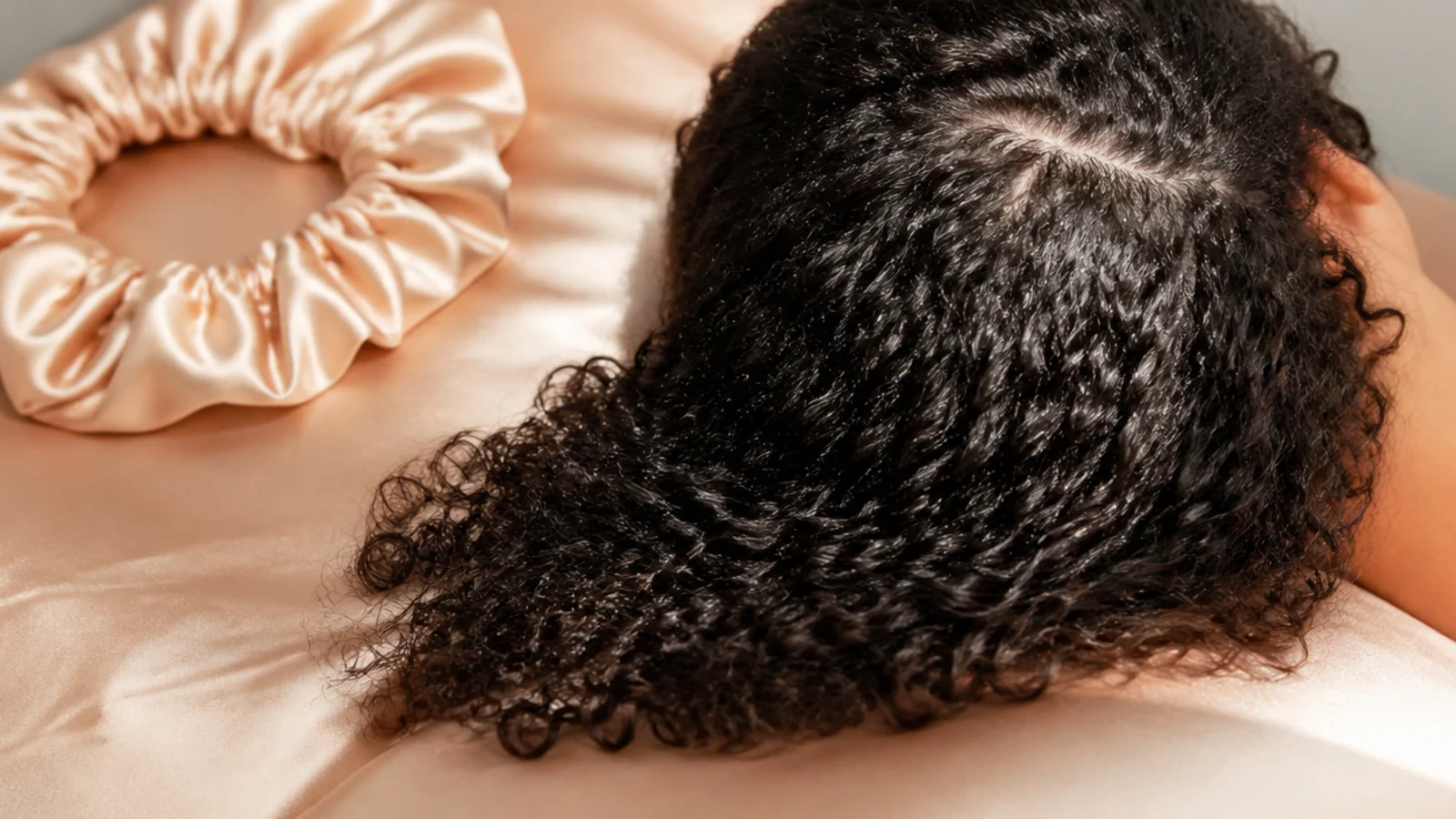Curly hair resting on a champagne satin pillowcase next to a satin sleep bonnet