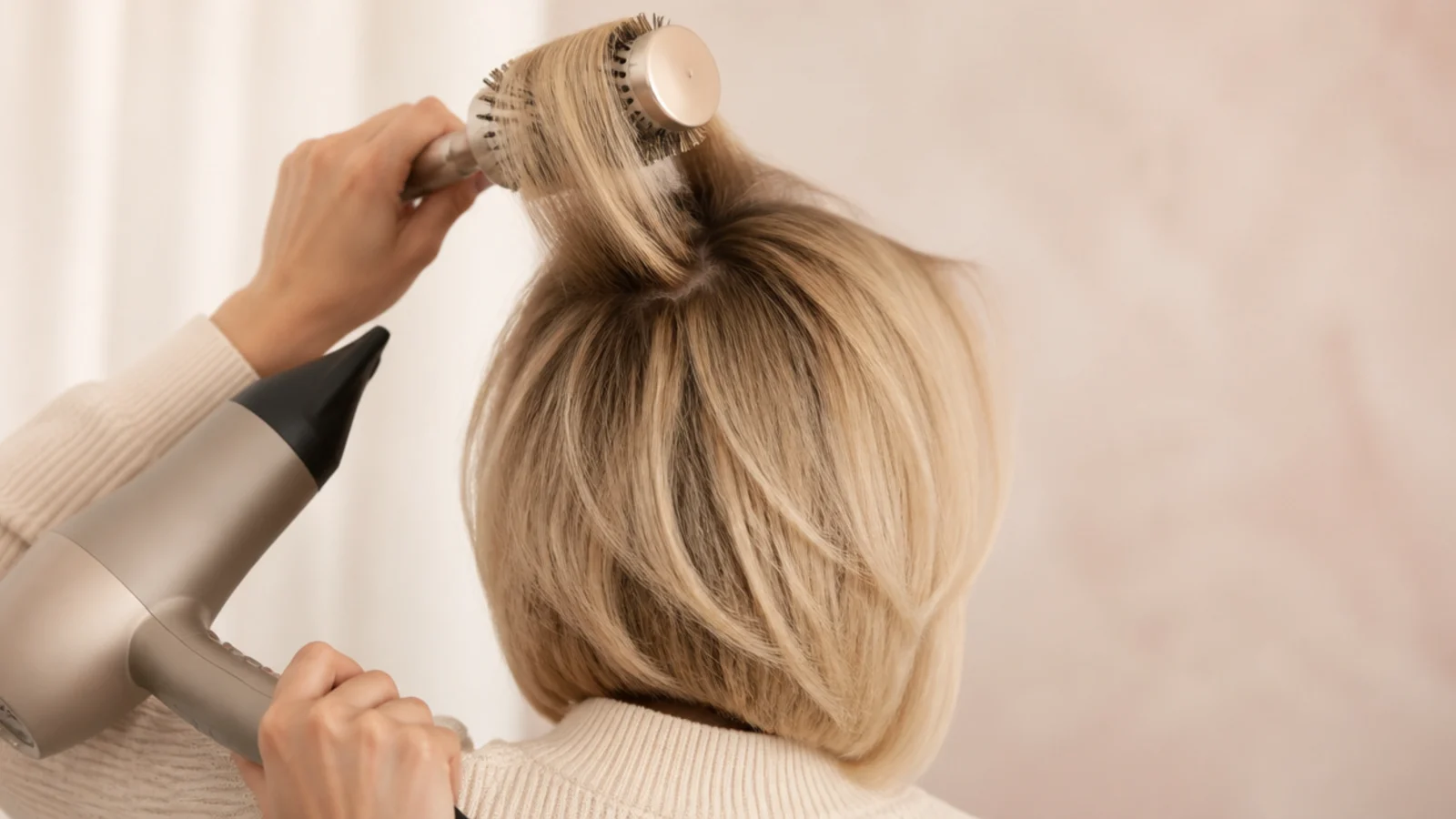 Large round brush lifting a section of a blonde bob at the root during a blow dry, from behind