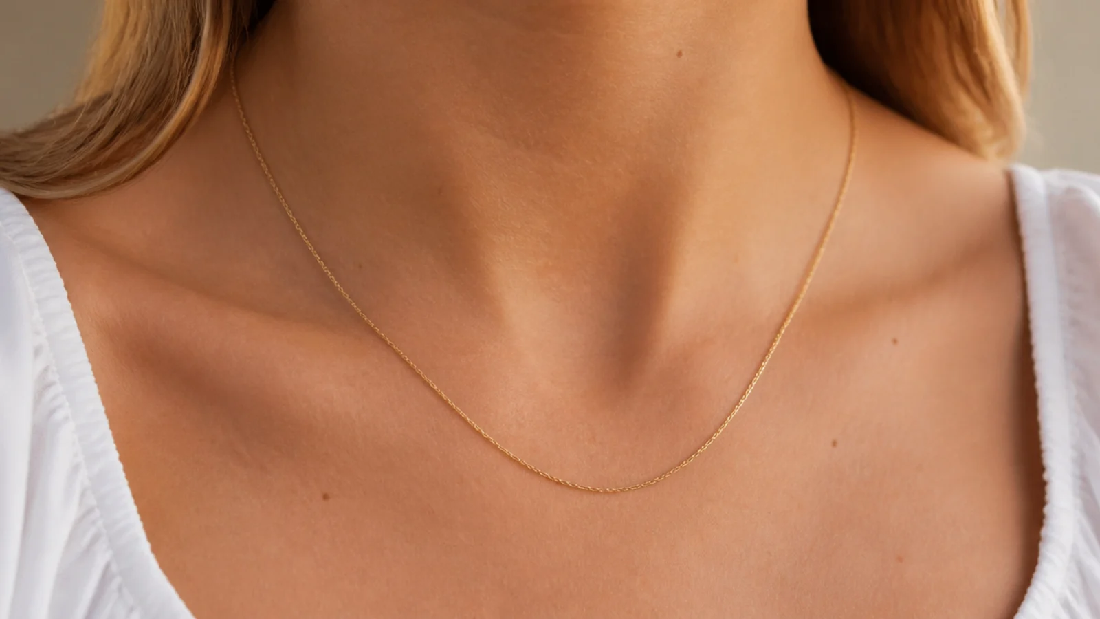 Faceless close-up of a thin gold chain necklace hitting just below the collarbone over a white top