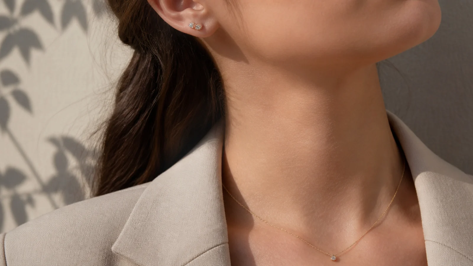 Faceless close-up of minimal interview jewelry with small studs and a thin chain over a structured jacket