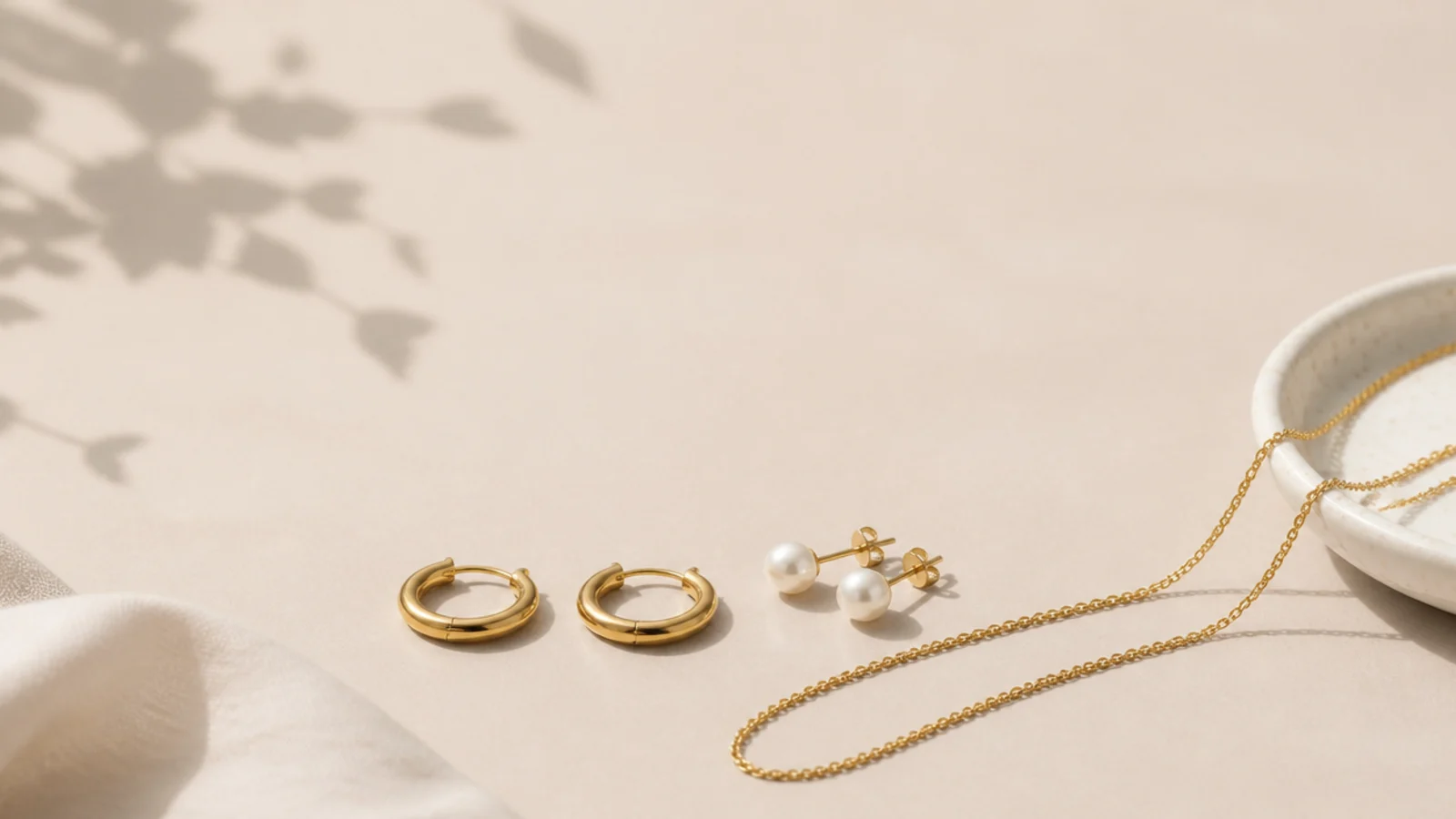 Faceless work jewelry flatlay with gold huggie hoops, a thin chain and pearl studs on a neutral desk