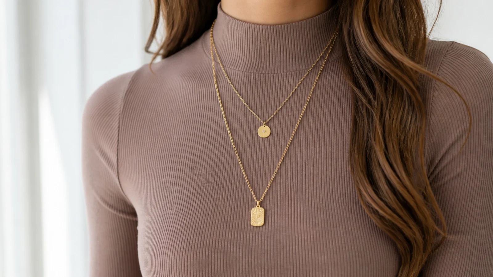 Faceless waist-up shot of two long gold chains layered over a fitted mock-neck top