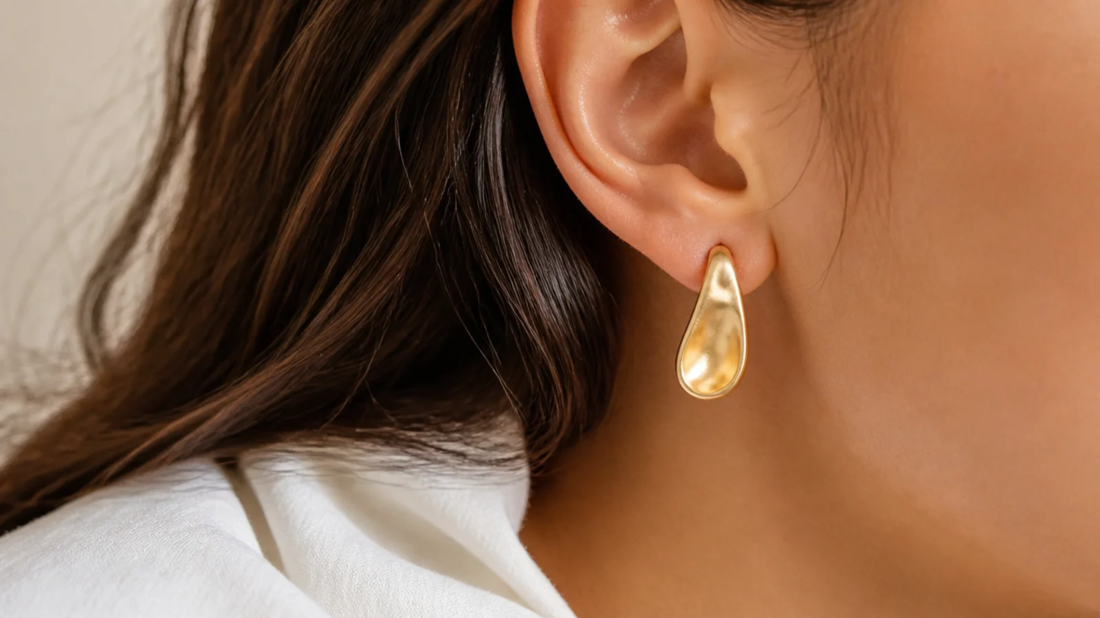 Faceless close-up of an organic molten-shaped matte gold drop earring below the earlobe