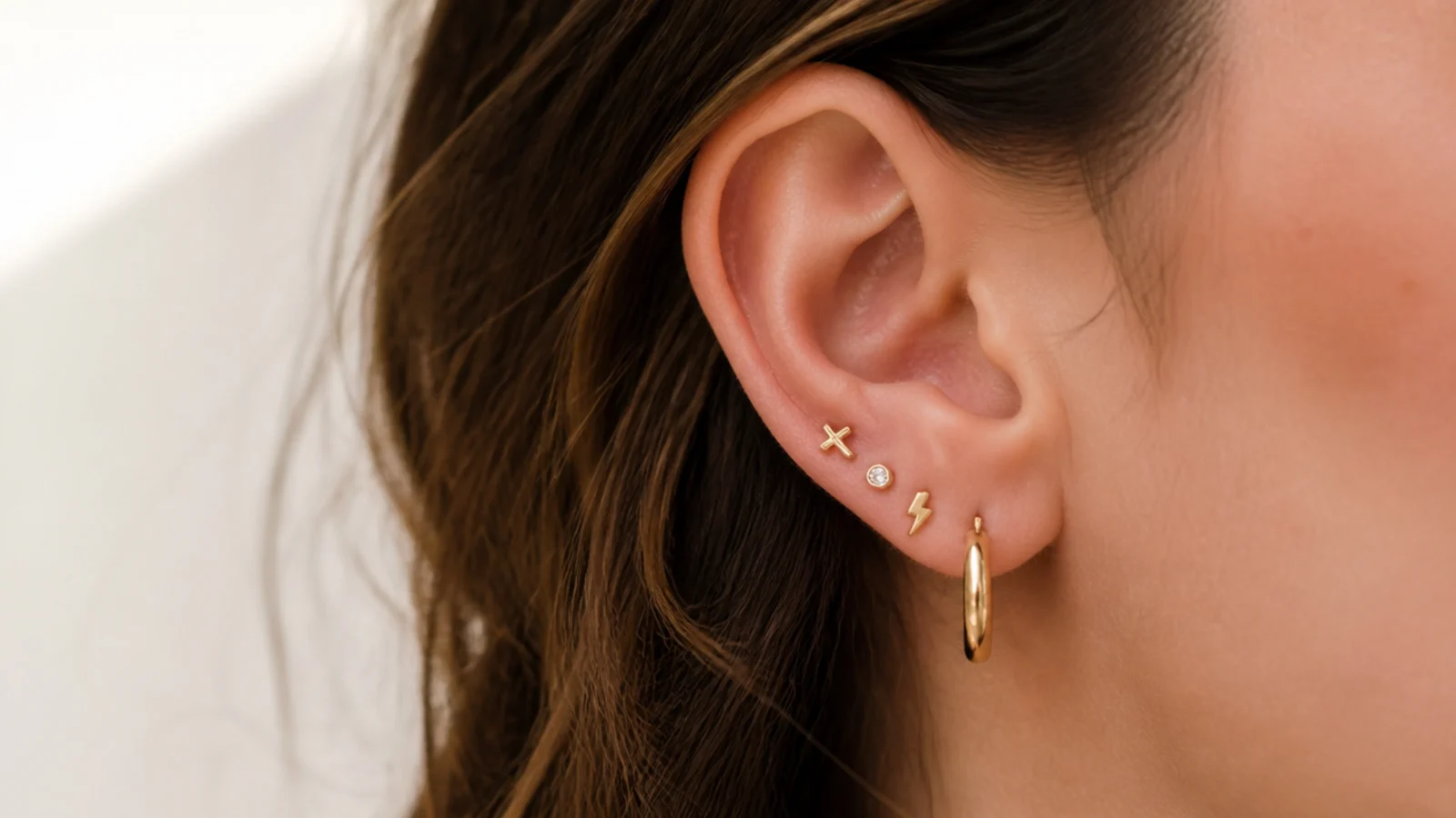 Faceless close-up of a curated ear stack with three tiny threaded gold studs and a small hoop
