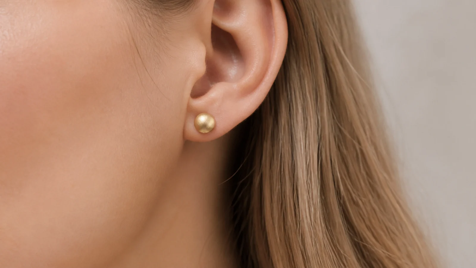 Faceless close-up of a small brushed matte gold dome stud earring on an earlobe