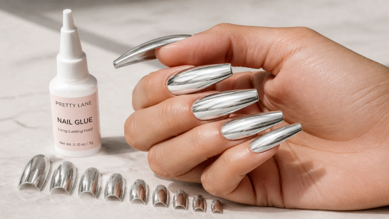 Long coffin press-on nails with a chrome finish arranged beside a bottle of nail glue