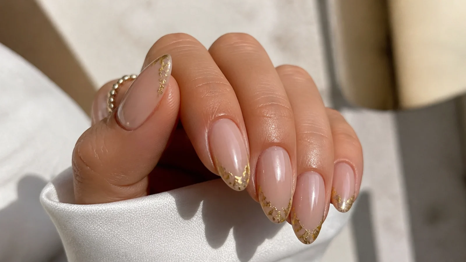 Almond nails with sheer nude bases and torn gold leaf fragments pressed along the French tip edge