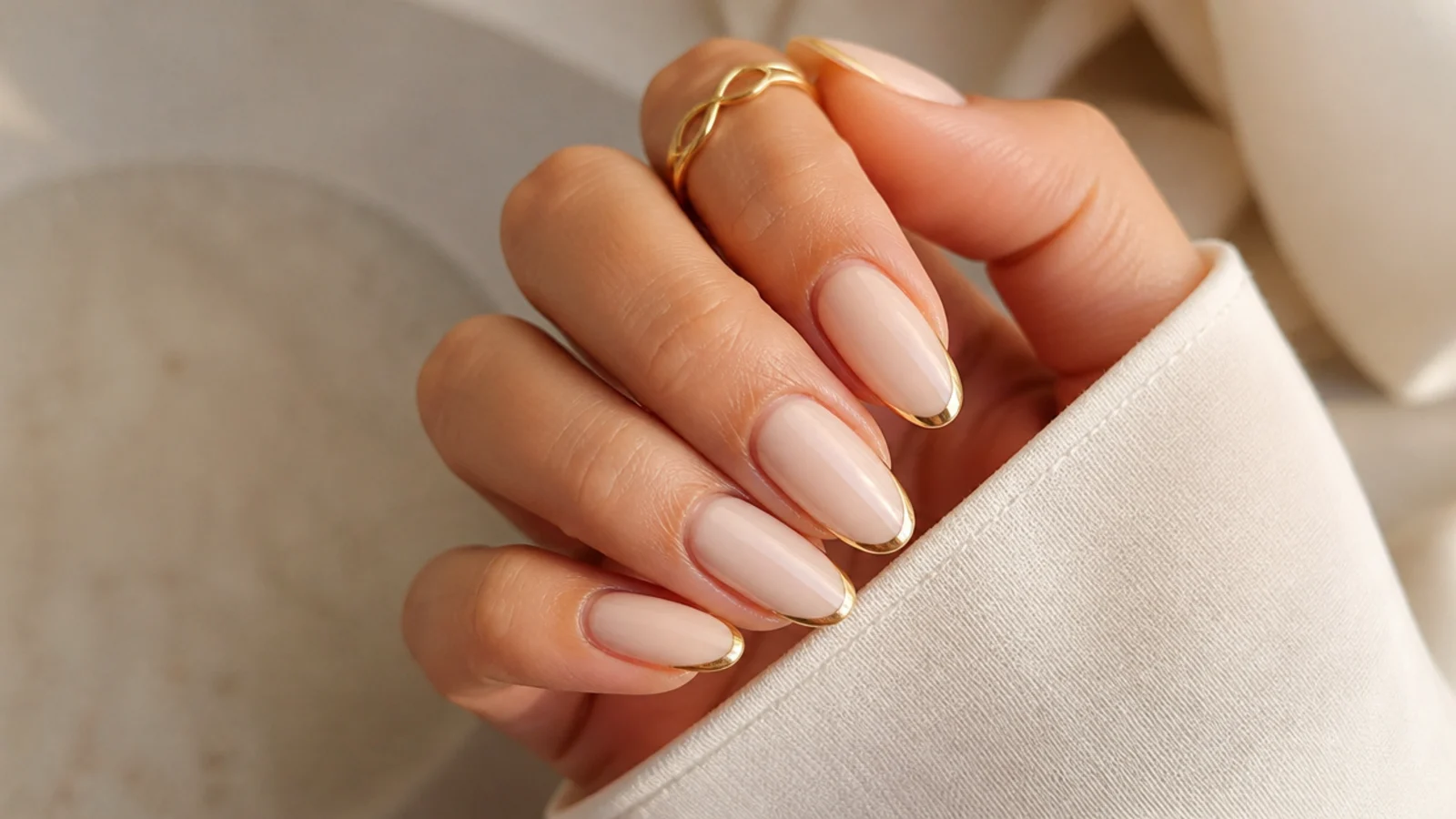 Milky beige almond nails outlined with a thin gold micro French line along the tip