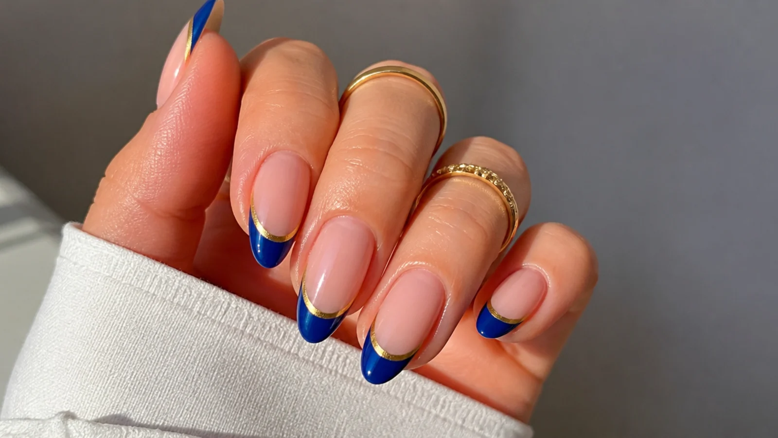 Nude nails with royal blue french tips outlined in thin gold on almond nails