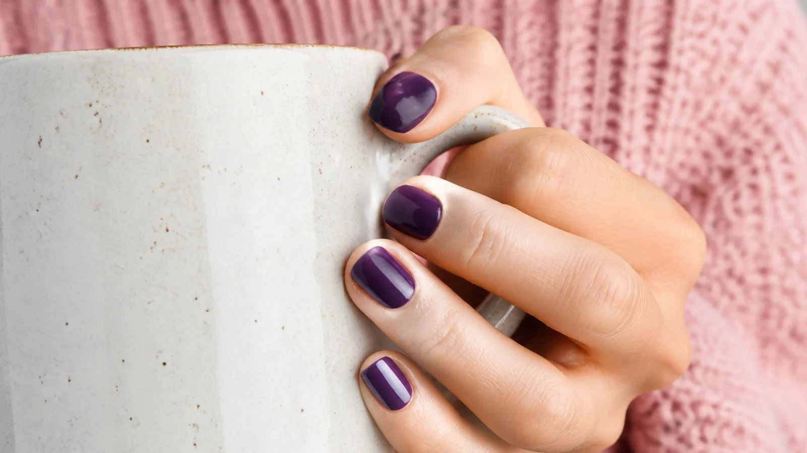 Short nails in deep plum purple cream gel with a glossy finish on a hand holding a mug
