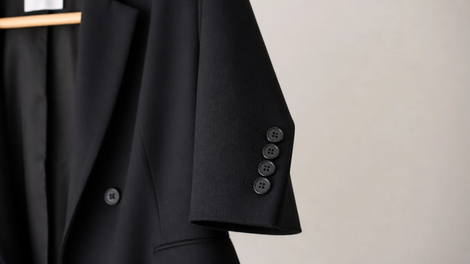 Faceless close-up of a matte black wool blazer sleeve and cuff on a hanger against a plain background