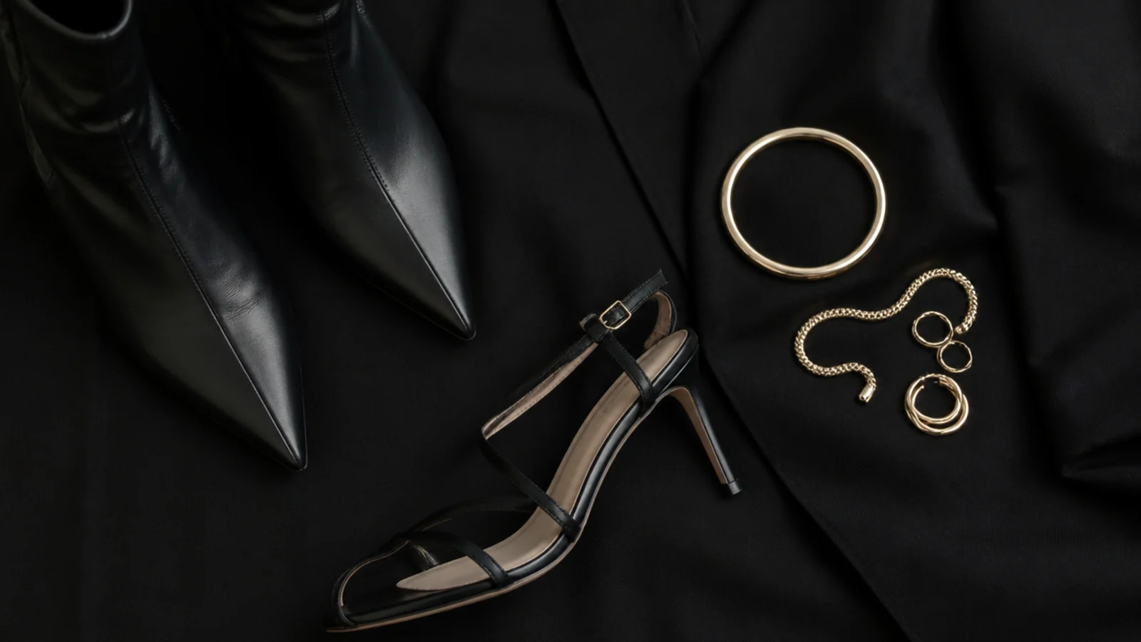 Faceless flat-lay of pointed black boots, a black strappy heel and stacked gold jewelry against black fabric