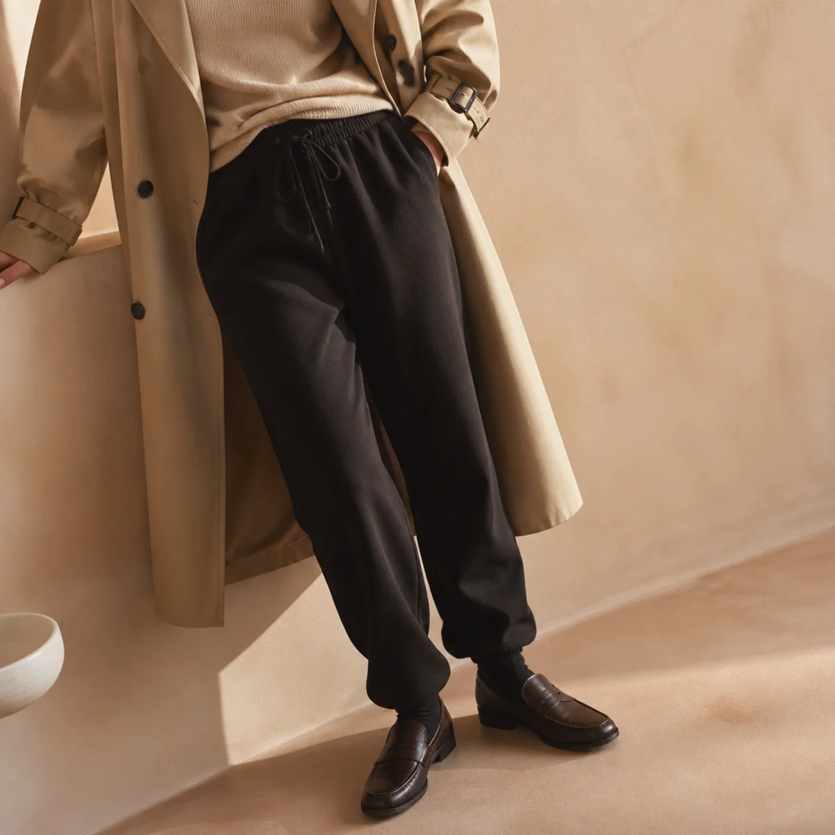 Faceless full-length shot of a beige trench coat worn open over black sweatpants and loafers