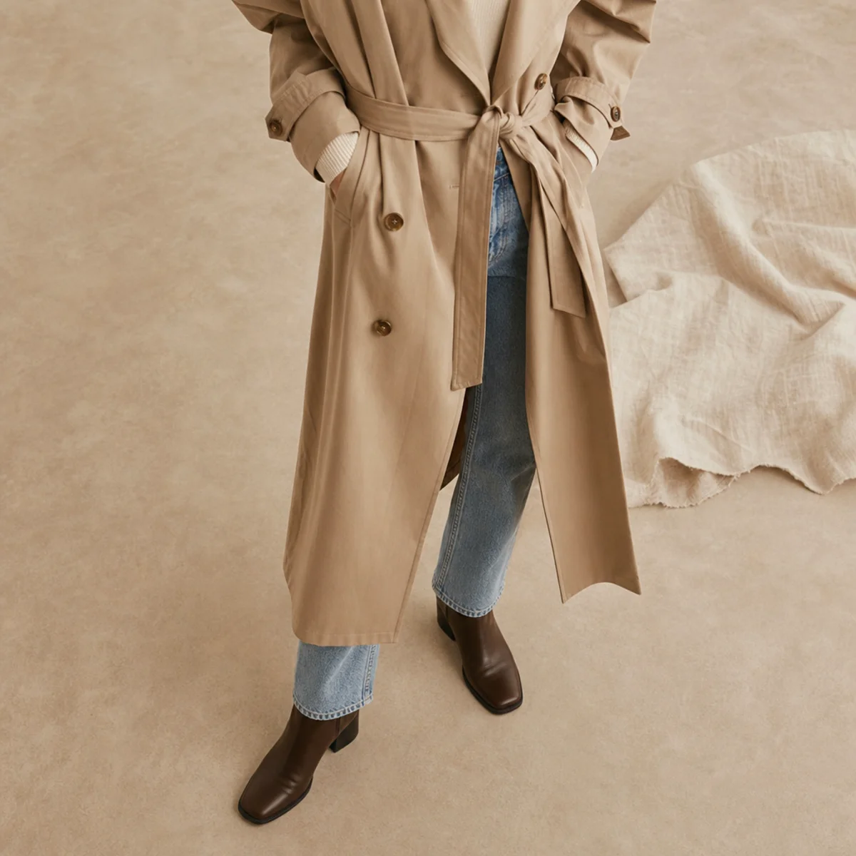 Faceless full-length shot of a belted beige trench over straight jeans with ankle boots